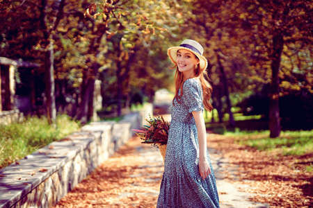 Glad redhead girl walks alone in autumn park on sunny warm day while holds handbag Indian summerの写真素材