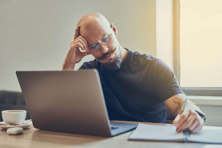 Adult caucasian bald man manager using laptop while writing notes in notebook at workplace.の写真素材