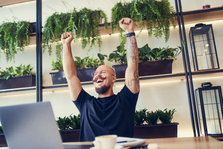 Happy young businessman sitting at laptop excited by good news online, lucky winner raising hand upの写真素材