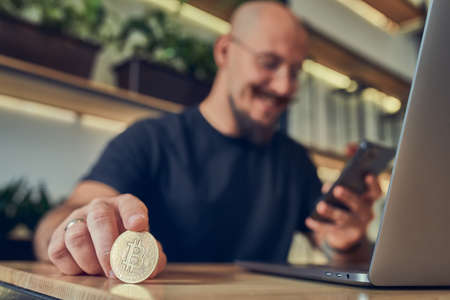 Successful bald businessman holds a gold bitcoin coin in his hand while screening smartphone.の写真素材