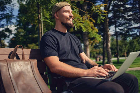 young caucasian freelancer sits on the bench in the park with laptop and enjoys relaxes free time