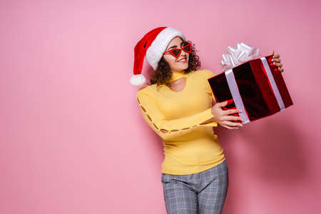 Smiling curly girl in Santas hat, sunglasses holds giftbox while stands on pink background New Year