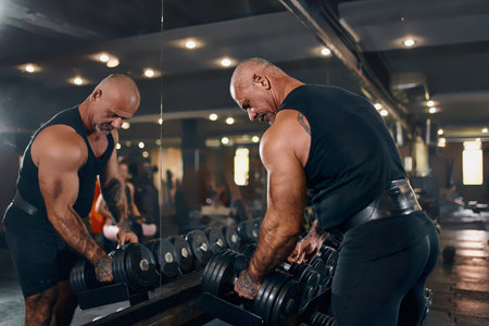 Bald male bodybuilder takes dumbbells in arms while posing in gym in dark. Concept of bodybuilding.の写真素材