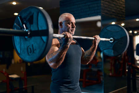 Older Bodybuilder athlete trains in the gym while pumping up biceps muscles. Sporty guy with barbellの写真素材
