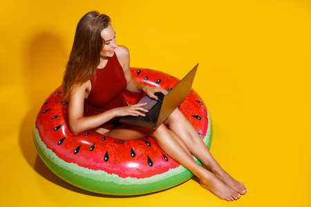 woman in swimsuit sits in inflatable ring works on laptop on yellow background. freelance. Travelの写真素材
