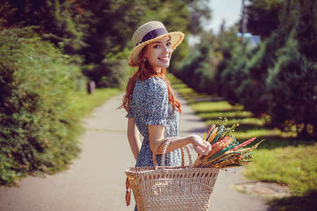 back view of young smiling adult girl with stylish straw bag and straw hat who walks in park outdoorの写真素材