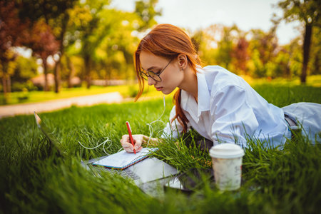 Female student prepares for exams using laptop makes notes lying on the grass. Education conceptの写真素材