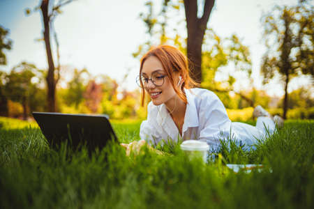 beautiful smiling girl student uses laptop pc while lying on the grass outdoors. Education conceptの写真素材