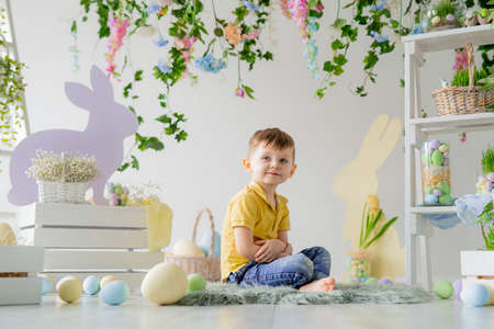 Little boy sits on wooden floor in Easter studio decoration with flowers, wooden rabbits, dyed eggsの写真素材