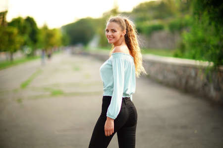 Back view of stylish smiling beauty girl who posing while wears in summer bright clothes in the park