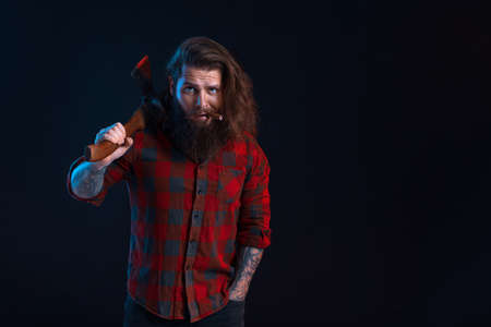 Emotional man with cigar holds old axe. Bearded lumberjack in checkered shirt with long hairの写真素材