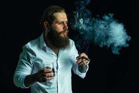 Handsome bearded man in white shirt blowing smoke holds glass of whiskey and cigar. Studio shotの写真素材