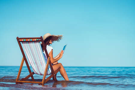 Back view of woman in sunhat who using laptop while lying on the beach chaise longue at the seasideの写真素材