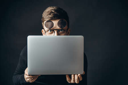 Man hiding behind laptop, looking at camera with big eyes, updating operation system. Indoor studio shot isolated on black backgroundの写真素材