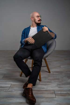 Hipster bearded man with moustache wearing glasses who sitting on chair in studio isolated grey background hold clipboard having thoughtful serious lookの写真素材