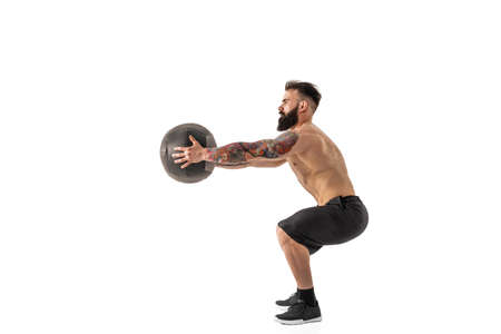 Muscular shirtless tattooed bearded male exercising fitness weights Medicine Ball in studio isolated...