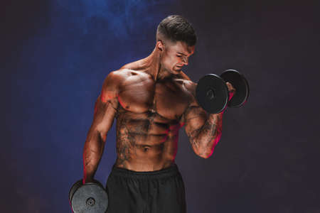 Brutal sweaty strong young man athlete with naked upper body standing doing workout with dumbbels...
