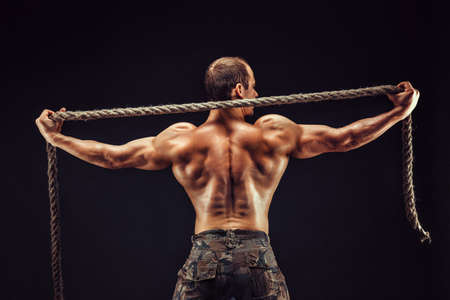Back view of handsome sexual strong young man, muscular body holding rope with hands standing posing...