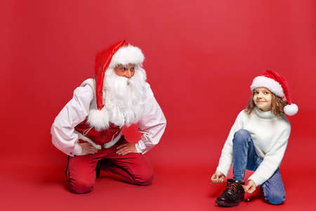 Cute little girl in Christmas hat trying on a boot with Santa against red studio background. Isolateの写真素材