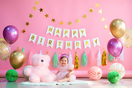Baby girl sits near paper balls and balloons with teddy in holiday dress on pink birthday backgroundの写真素材