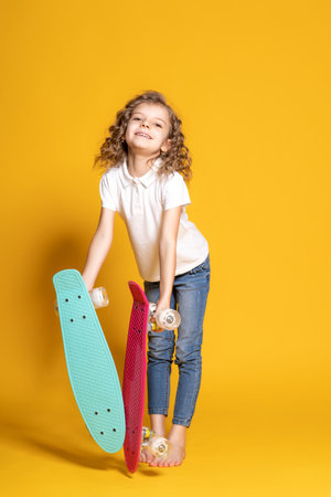 Full length of little girl in white polo, blue jeans who holds two skateboards on yellow backgroundの写真素材