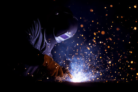 Welder engages in welding process, sparks erupting from welding torch in dimly lit workshop. Creative metalworking setting, highlighting skill and precision.の素材
