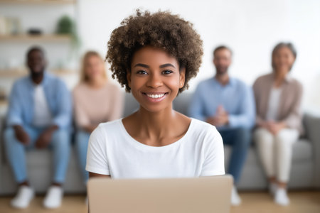 Woman smiling at camera while using laptop, engaged in work at home office. Comfortable modern setting with friends in the background. Concept of teamwork, remote work, friendship.の素材
