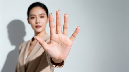 Woman confidently holds hand up in stop gesture with slight smile. Clean and bright background enhances focus on her expression. Concept of communication, professionalism, self-assurance.の素材