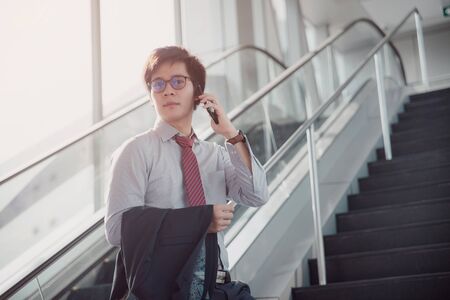 Handsome smiling confident businessman talking something on smart phone and walking inside in airport. Casual young businessman wearing white shirt with red tie and holding a black suit.の写真素材