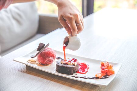 Human hand who try to drop strawberry jam from a little white jar. Close up the chocolate lava dessert with strawberry ice cream on white plate.の写真素材