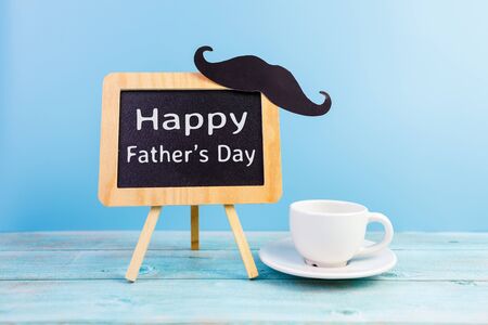 Happy Father's day concept. A black mustache paper, a cup of coffee and the text written that Happy Father's Day on blackboard isolated on blue background.の写真素材