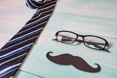 Happy Father's day concept. A black mustache paper, a blue necktie and glasses on wooden table.