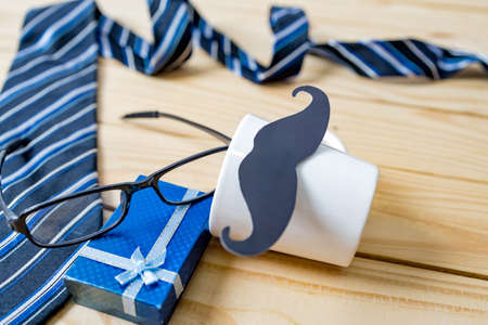 Happy Father's day concept. A black mustache paper, a blue necktie, a blue gift box and glasses on wooden table.の写真素材