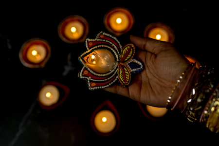 Happy Diwali - Woman hands with henna holding lit candle isolated on dark background. Clay Diya lamps lit during Dipavali, Hindu festival of lights celebration. Copy space for text.の写真素材