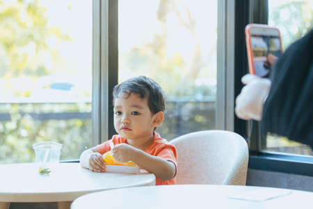 Asian boy enjoying delicious orange cake in a restaurant with a smartphone secretly taking picturesの写真素材