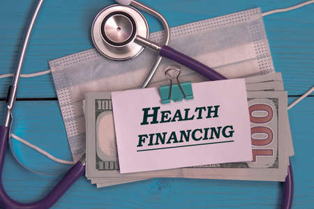 HEALTH CARE FINANCING - words on white sheet against background of banknotes, medical mask and stethoscope. Medical and finance conceptの写真素材