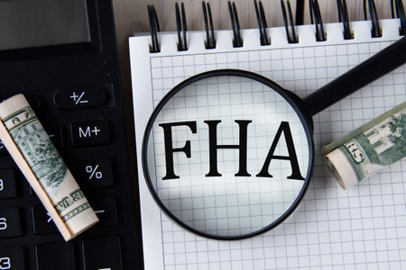 Letters FHA (Federal Housing Administration) under a magnifying glass on a white notebook against the background of a fragment of a calculator and banknotesの写真素材