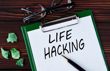 LIFE HACKING - words on a white sheet on a brown wooden background with a pen and glassesの写真素材