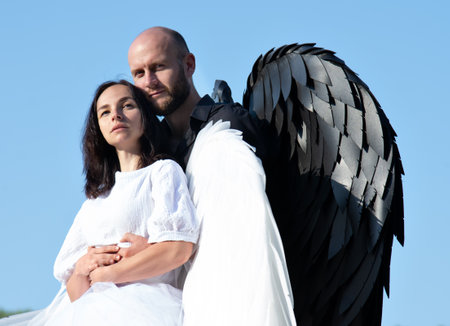 Portrait of a girl in a white dress with white wings and a man with black wings. Couple standing in an embrace against the background of a blue skyの写真素材