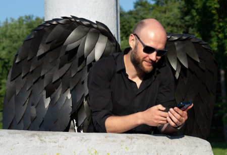 a handsome strong modern man in an angel costume with black wings against a background of light columns. Black angel in the modern world with a phone in his handsの写真素材