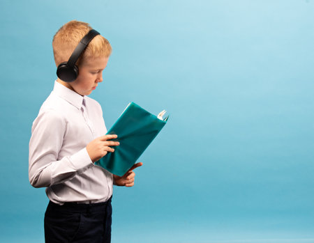 Little schoolboy with headphones on his head and a notebook on a blue background. Studio shot....