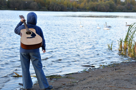 A person with a guitar looks at a lake with ducks. Rear view.の写真素材