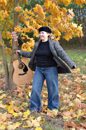 A girl with a guitar happily plays under a tree with fallen maple leaves. Beautiful autumn