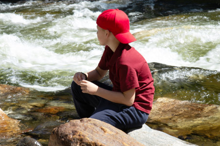 A boy enjoys a beautiful view of the forest, river, and mountains. The boy sits on a large rock on the bank of a mountain river.の写真素材