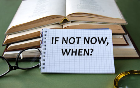 Notebook with IF NOW, WHEN? text on a green background with books and glasses. Info conceptの写真素材