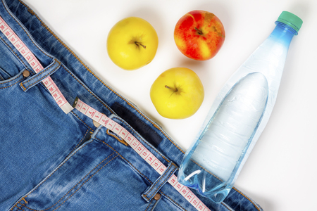 Healthy lifestyle concept. Jeans with a measuring tape instead of a belt, apples and a bottle of waterの写真素材