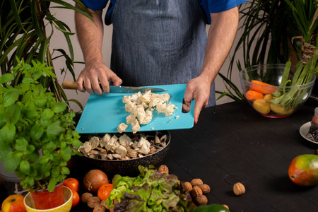 A man is cutting food on a blue cutting board. The food includes vegetables and nuts. The man is...