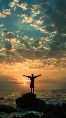 A man stands on a rock overlooking the ocean at sunset. He is holding his arms out in the air, as if he is celebrating or expressing joy. Concept of freedom and happinessの素材