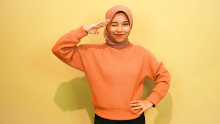Hijab woman saluting with hand on orange wall background.