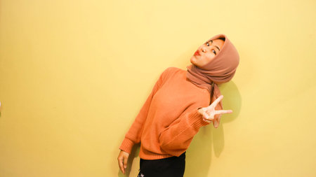 Hijab Asian woman show peace sign with orange wall background.の写真素材